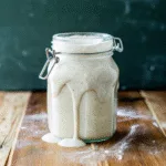 Overflowing, bubbly sourdough starter in a glass jar on a wooden surface, showing strong fermentation activity.